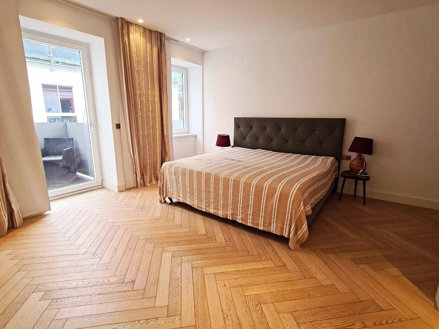 Schlafzimmer mit Fischgrätparkett, großem Bett und direktem Zugang zu einem kleinen Balkon.