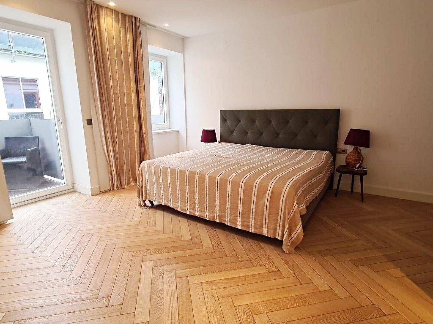 Helles Schlafzimmer mit elegantem Fischgrätparkett, großem Bett und Zugang zum Außenbereich.