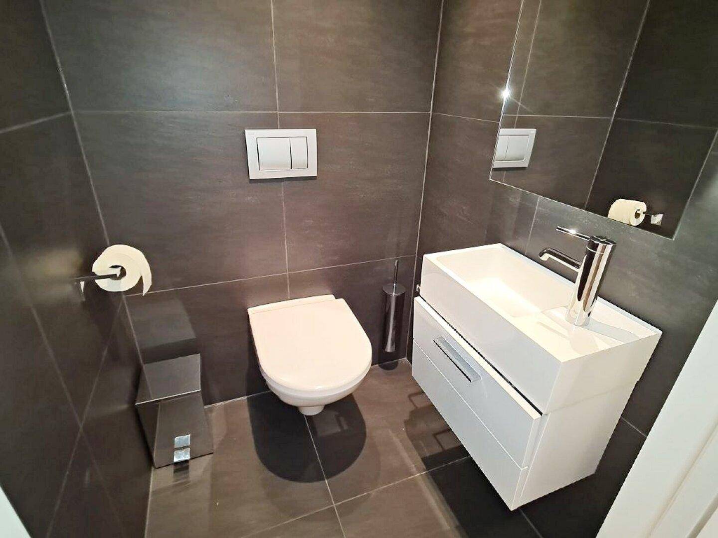 Modernes Gäste-WC mit dunklen Fliesen, wandhängender Toilette und weißem Waschtisch.