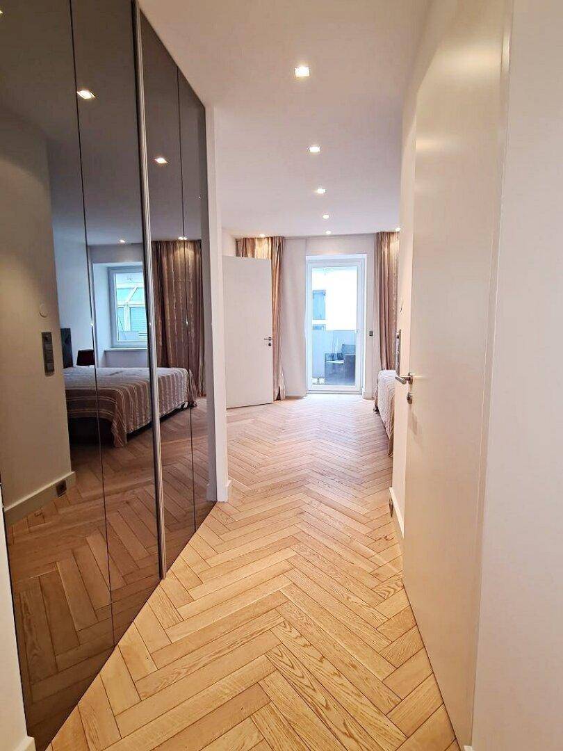 Heller Flur mit Fischgrätparkett, Spiegelschränken und Blick auf ein Schlafzimmer mit Balkonzugang.