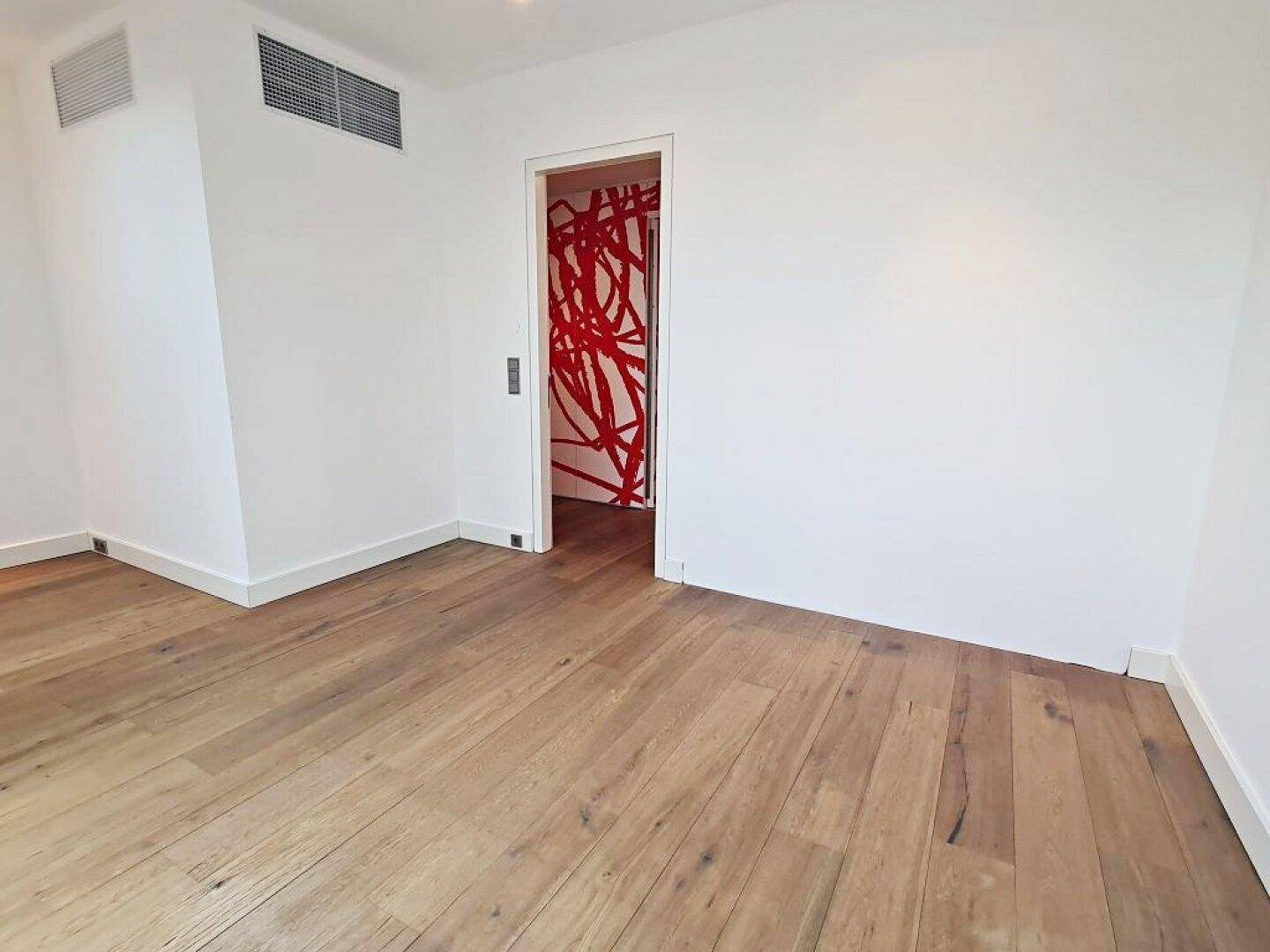 Leerer Raum mit Holzboden und weiß gestrichenen Wänden, angrenzend an einen Flur mit roter Wandkunst.