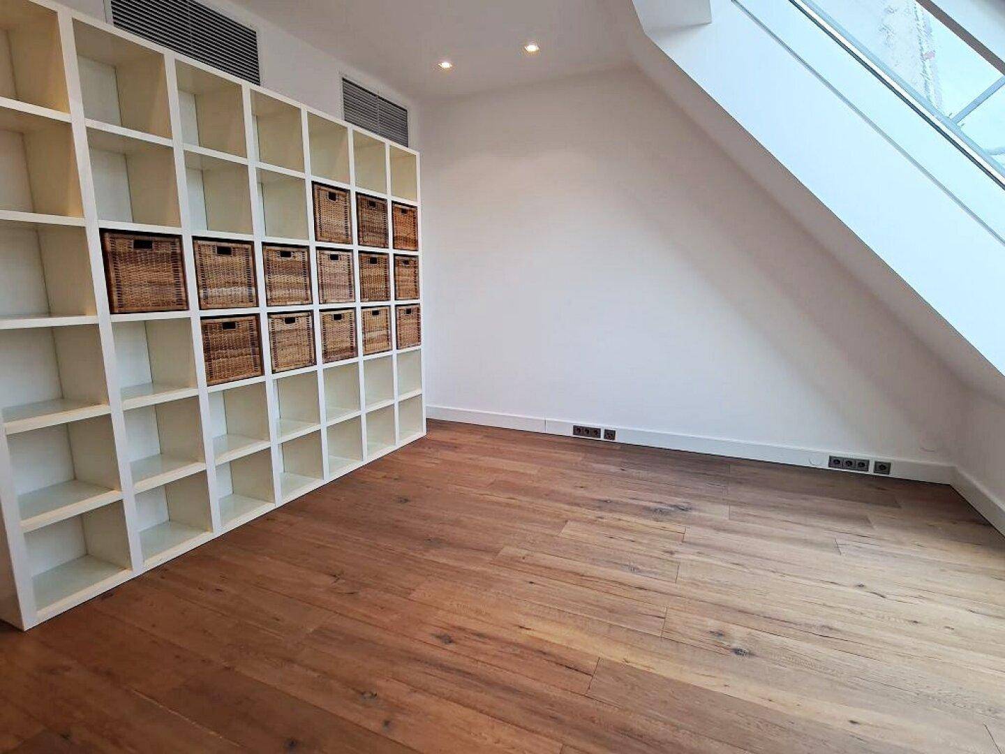Geräumiger Raum mit Holzboden, Dachschräge, Dachfenster und einem Regalsystem mit Körben.