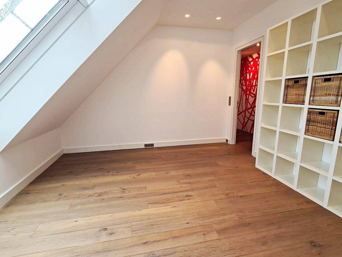 Heller Raum mit Holzboden, Dachschräge, Dachfenster und Regalsystem, angrenzend an einen farbigen Flur.