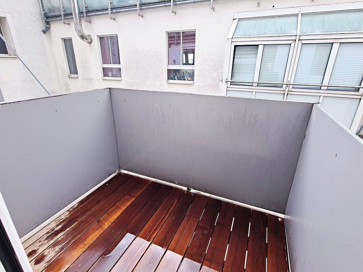 Kleiner Balkon mit dunklem Holzdeck und grauen Wänden, umgeben von Gebäudefassaden.