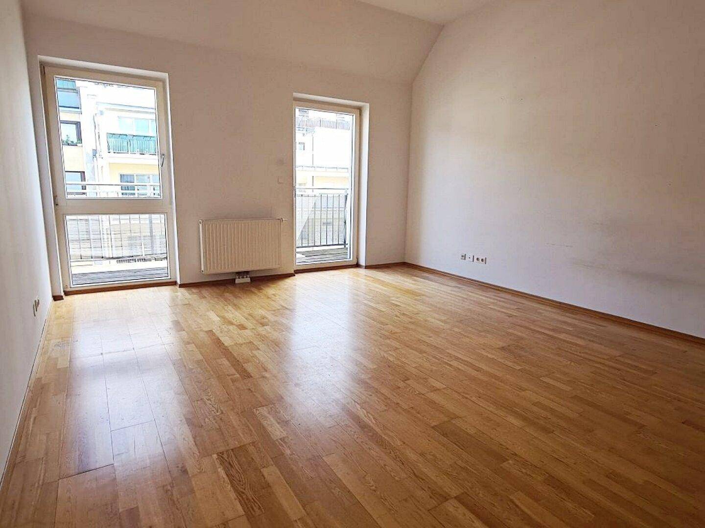 Geräumiges Wohnzimmer mit hellem Parkettboden und Zugang zu einem kleinen Balkon.