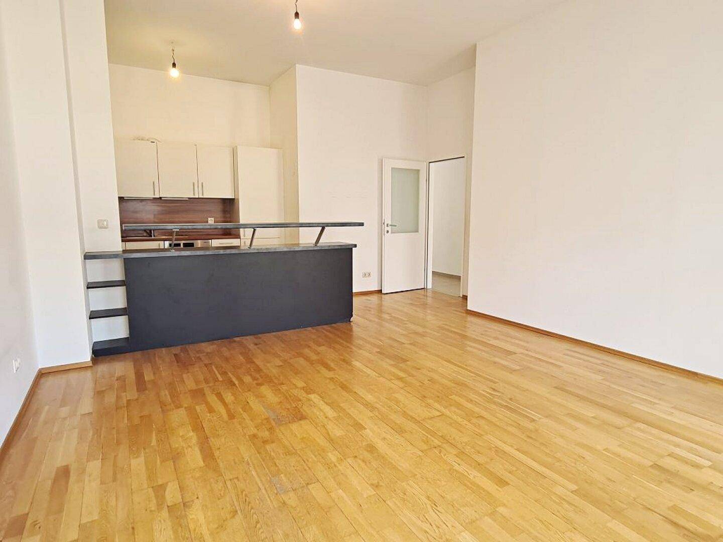 Offener Wohnbereich mit Parkett und integrierter Küchenzeile im Hintergrund, sehr hell.