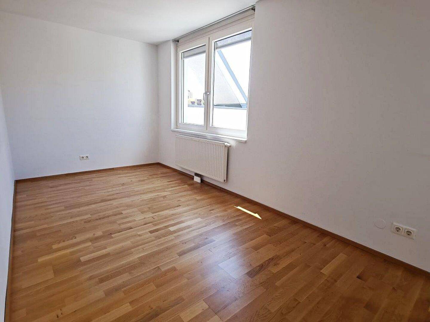 Helles Zimmer mit Parkettboden und Fenster mit Jalousien, bietet viel Licht.