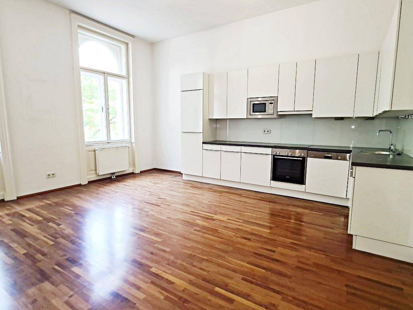 Geräumiger Wohnbereich mit integrierter weißer Küche, Parkettboden und hohen Fenstern.