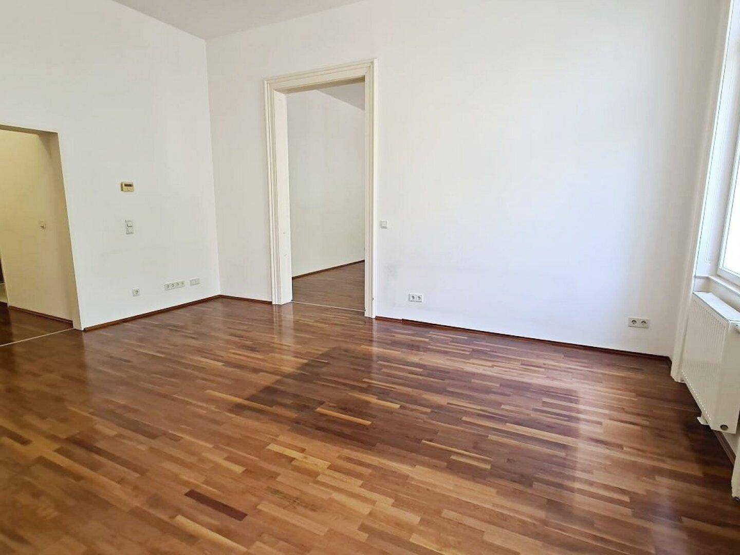 Geräumiges Wohnzimmer mit Parkettboden und hohen Decken, offen zu weiteren Räumen.