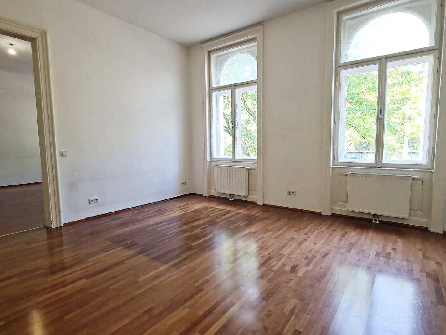 Helles Wohnzimmer mit Parkettboden, hohen Decken und zwei großen Bogenfenstern.