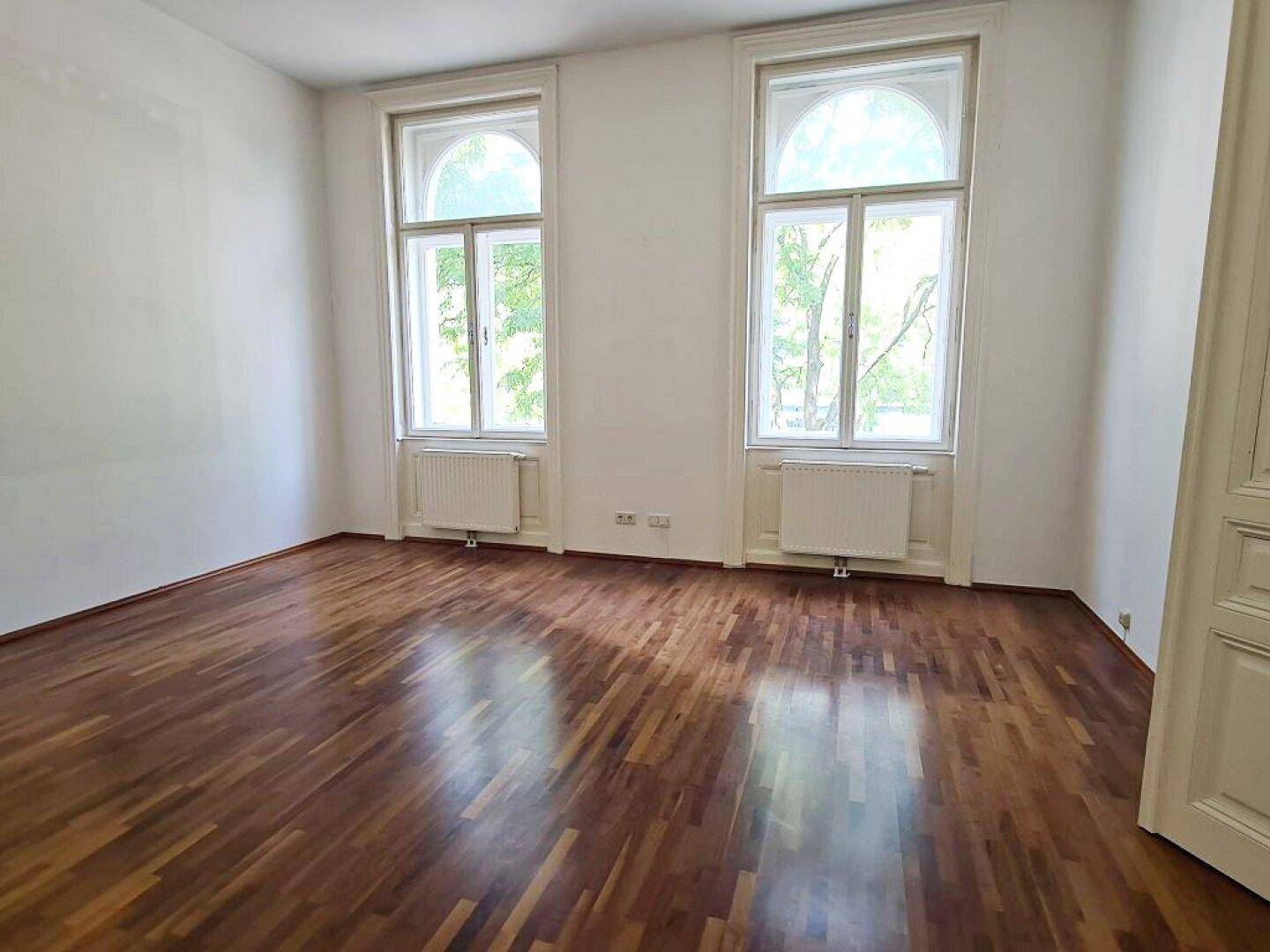 Helles Zimmer mit Parkettboden, hohen Decken und zwei großen Bogenfenstern.
