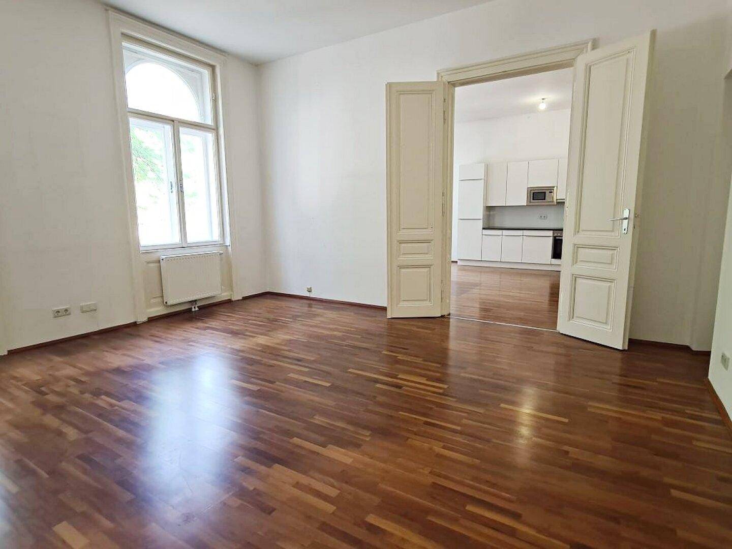 Helles Zimmer mit Parkettboden und einem großen Bogenfenster, mit Zugang zur Küche.
