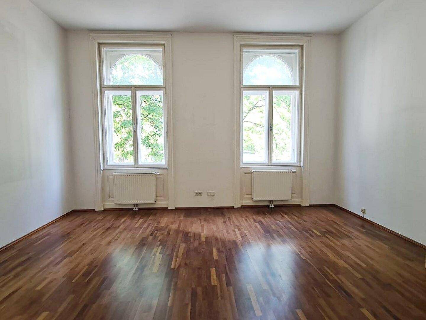 Helles Zimmer mit Parkettboden, hohen Decken und zwei großen Bogenfenstern.