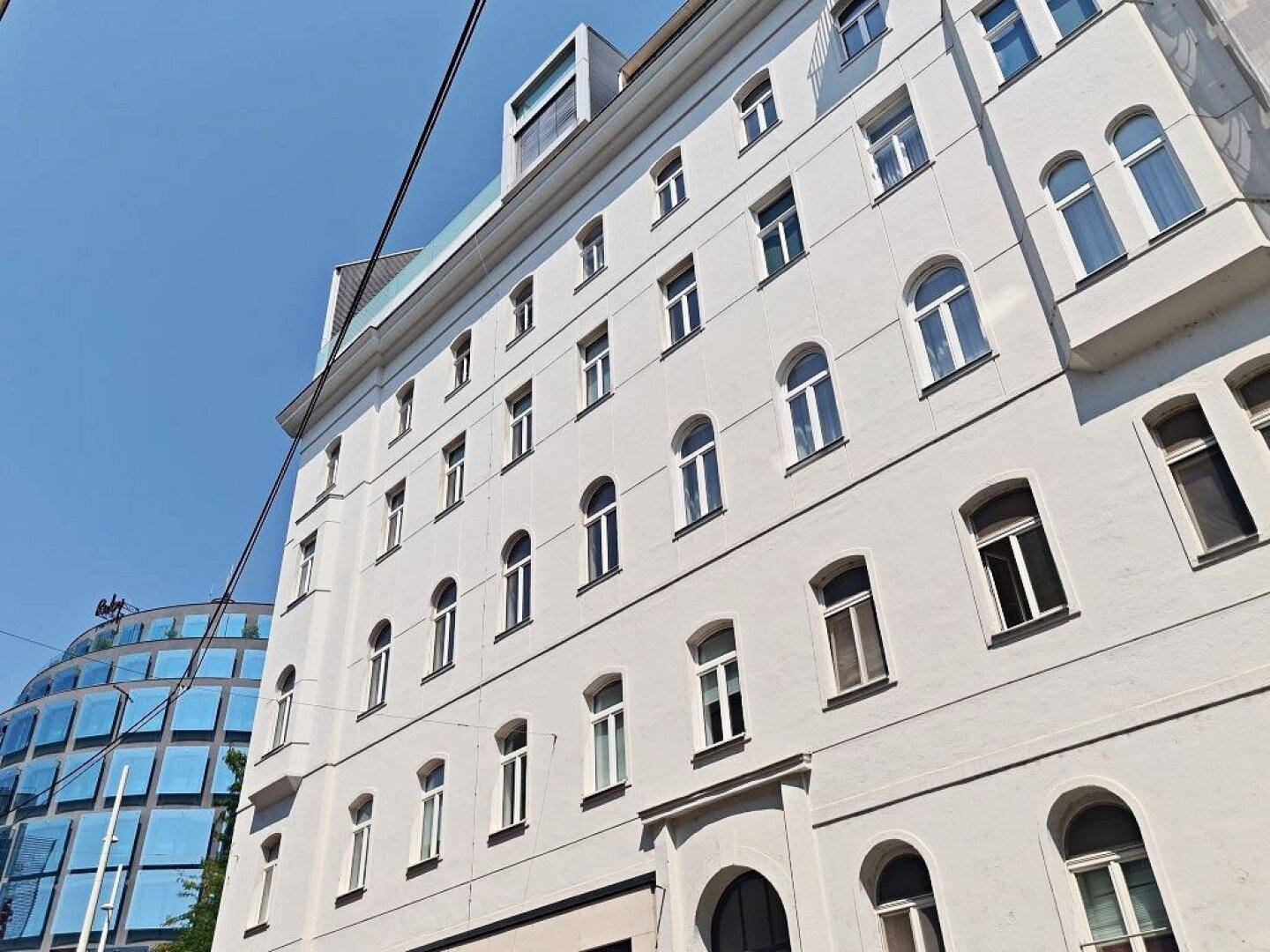Weiße Fassade eines Altbaus mit modernen Dachaufbauten und vielen Fenstern unter blauem Himmel.