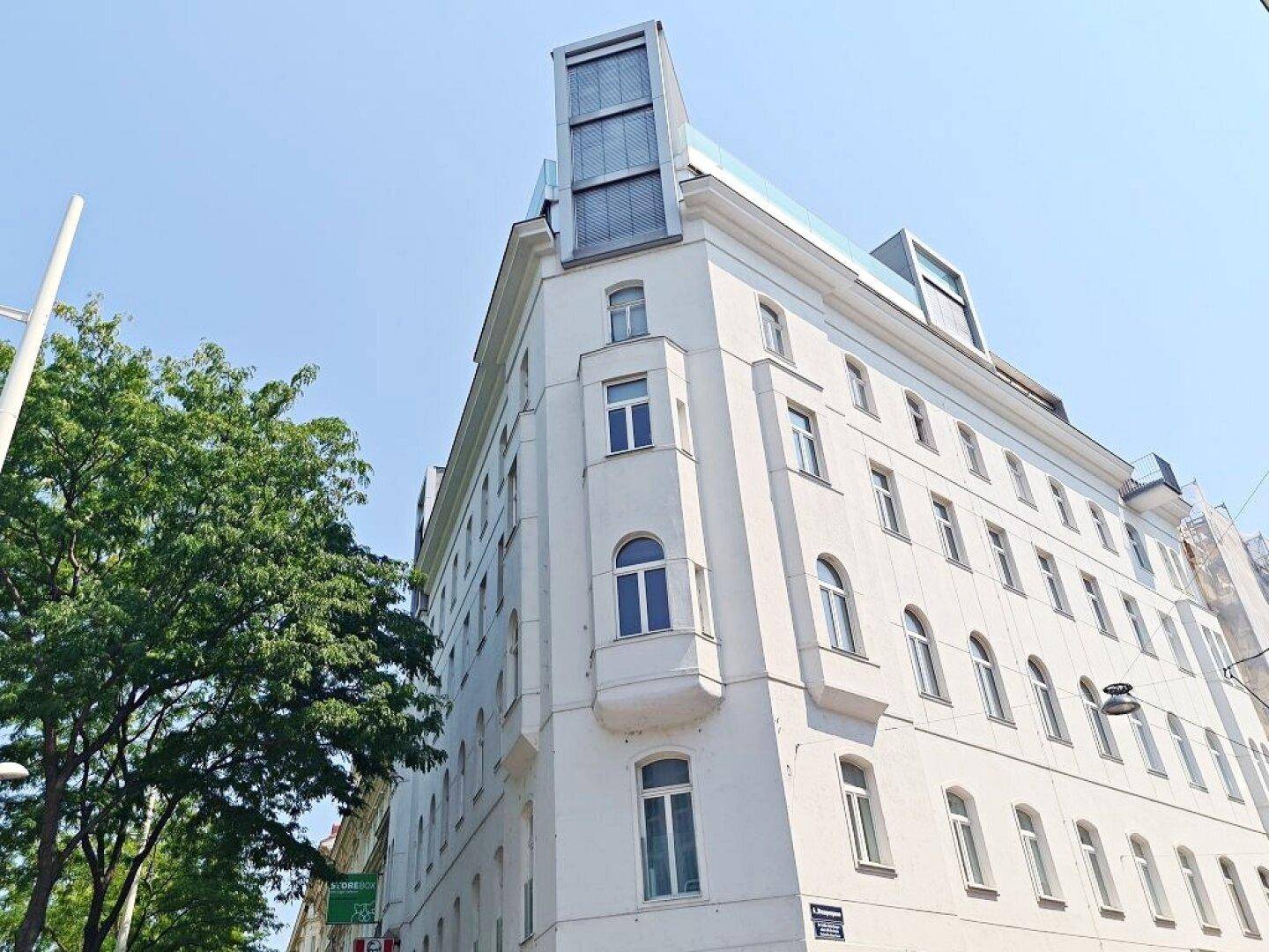 Eckansicht des Gebäudes mit weißer Fassade, modernen Dachaufbauten und einem Baum im Vordergrund.