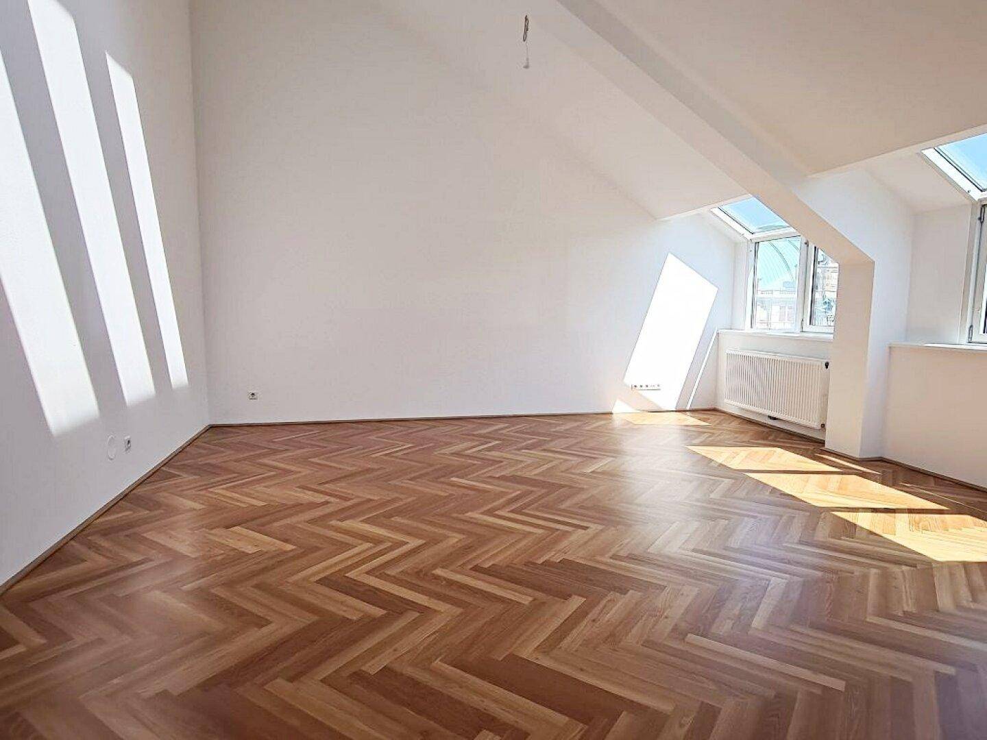 Heller Wohnraum mit Fischgrätparkett und schrägen Wänden, durchflutet von Sonnenlicht durch die Fenster.