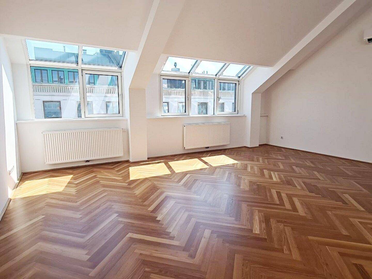 Heller Wohnbereich mit Fischgrätparkett, Dachschrägen und großen Fenstern, die viel Tageslicht hereinlassen.