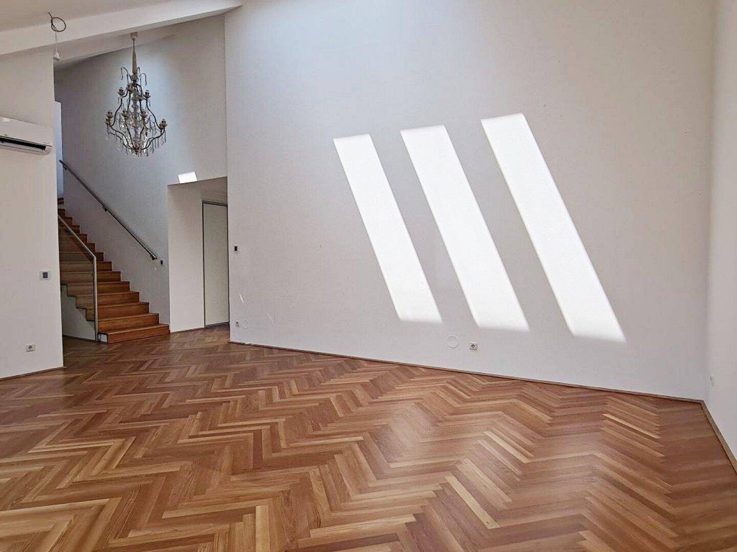 Heller Wohnraum mit Fischgrätparkett und schrägen Wänden, durchflutet von Sonnenlicht durch die Fenster.