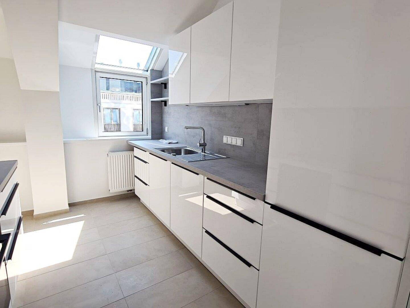 Moderne Küche mit weißen Hochglanzschränken, grauer Arbeitsplatte und Dachfenster für natürliches Licht.