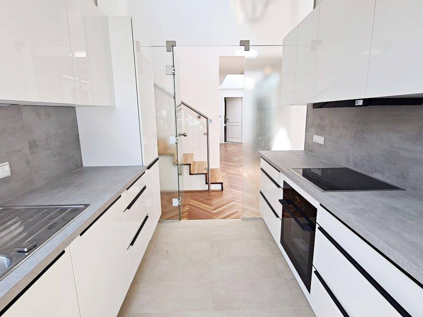 Moderne Küche mit weißen Hochglanzschränken und Blick auf den Flur mit Treppe durch eine Glaswand.