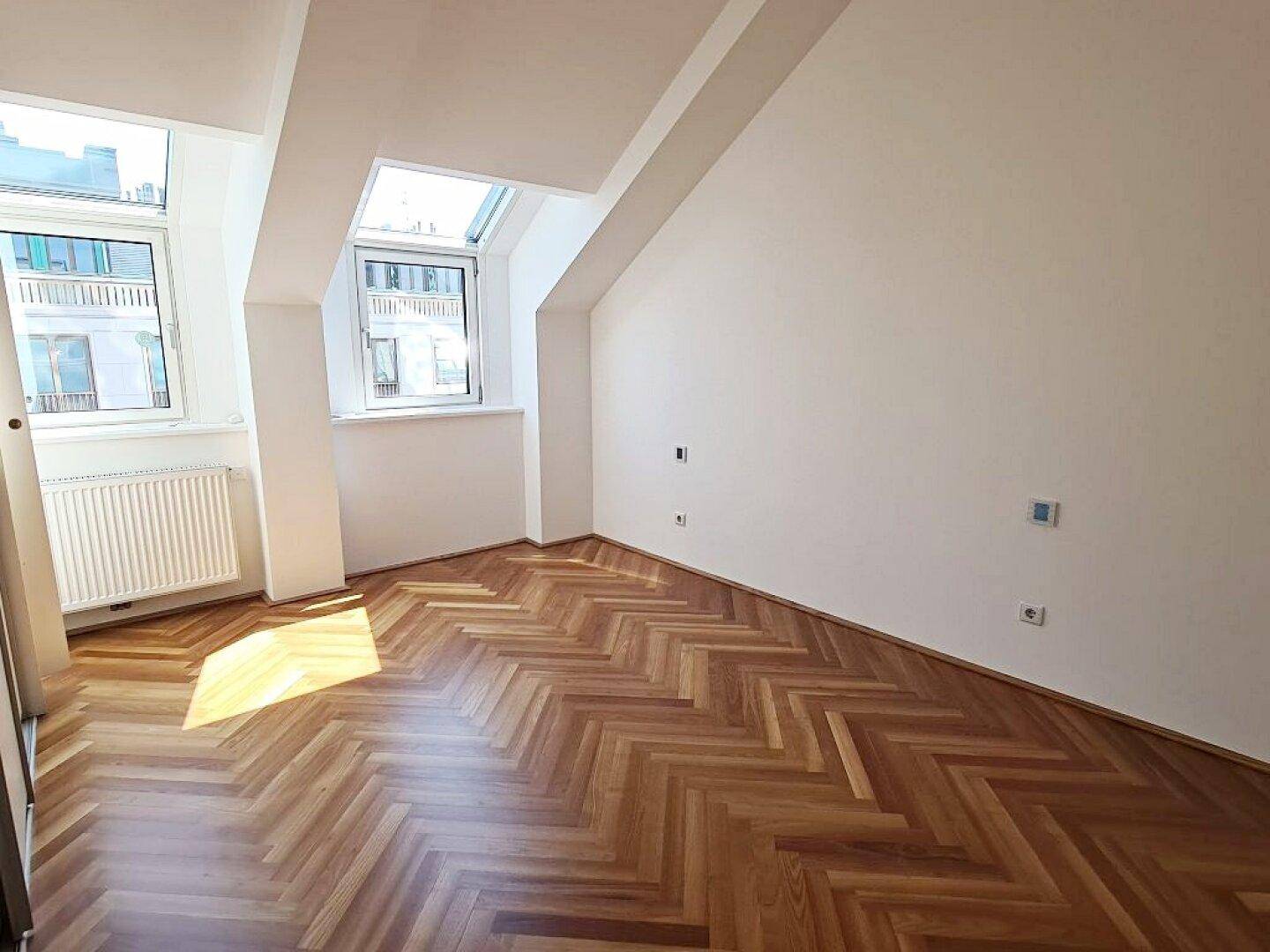 Heller Wohnbereich mit Fischgrätparkett, Dachschrägen und großen Fenstern, die viel Tageslicht hereinlassen.