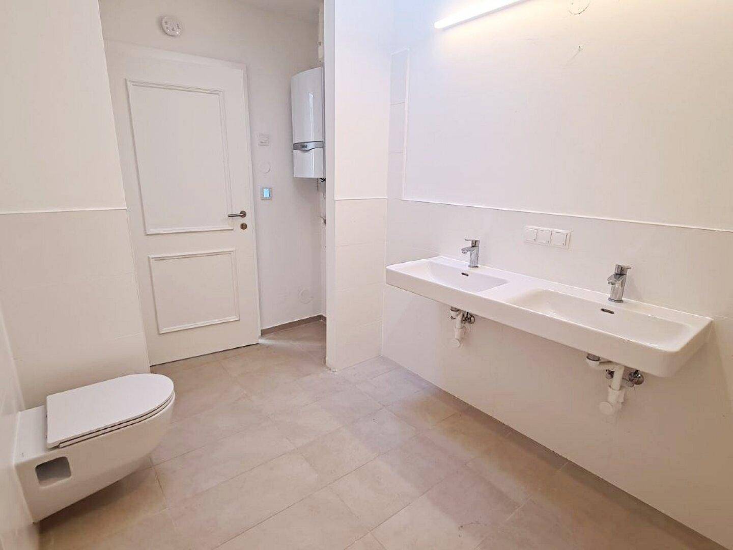 Helles Badezimmer mit wandhängendem WC, Doppelwaschbecken und einem Warmwasserbereiter an der Wand.