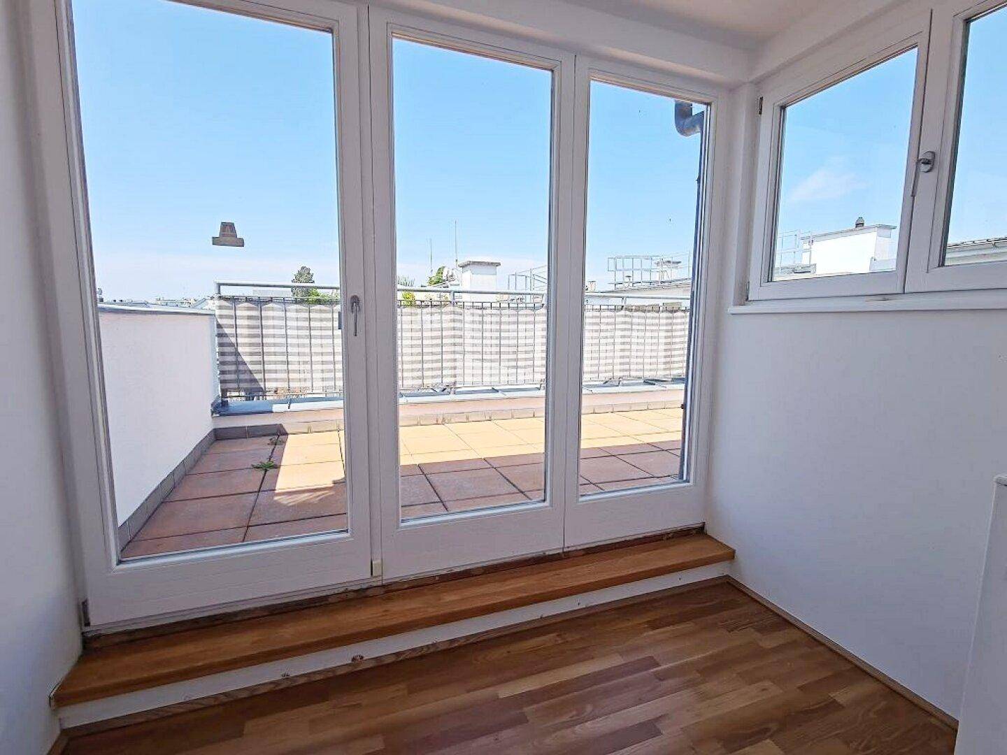 Heller Innenraum mit großen Glastüren, die zu einer sonnigen Dachterrasse mit Fliesenboden führen.