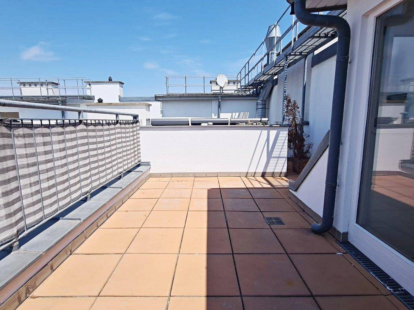 Weitläufige Dachterrasse mit Fliesenboden und Blick über die umliegenden Dächer unter blauem Himmel.