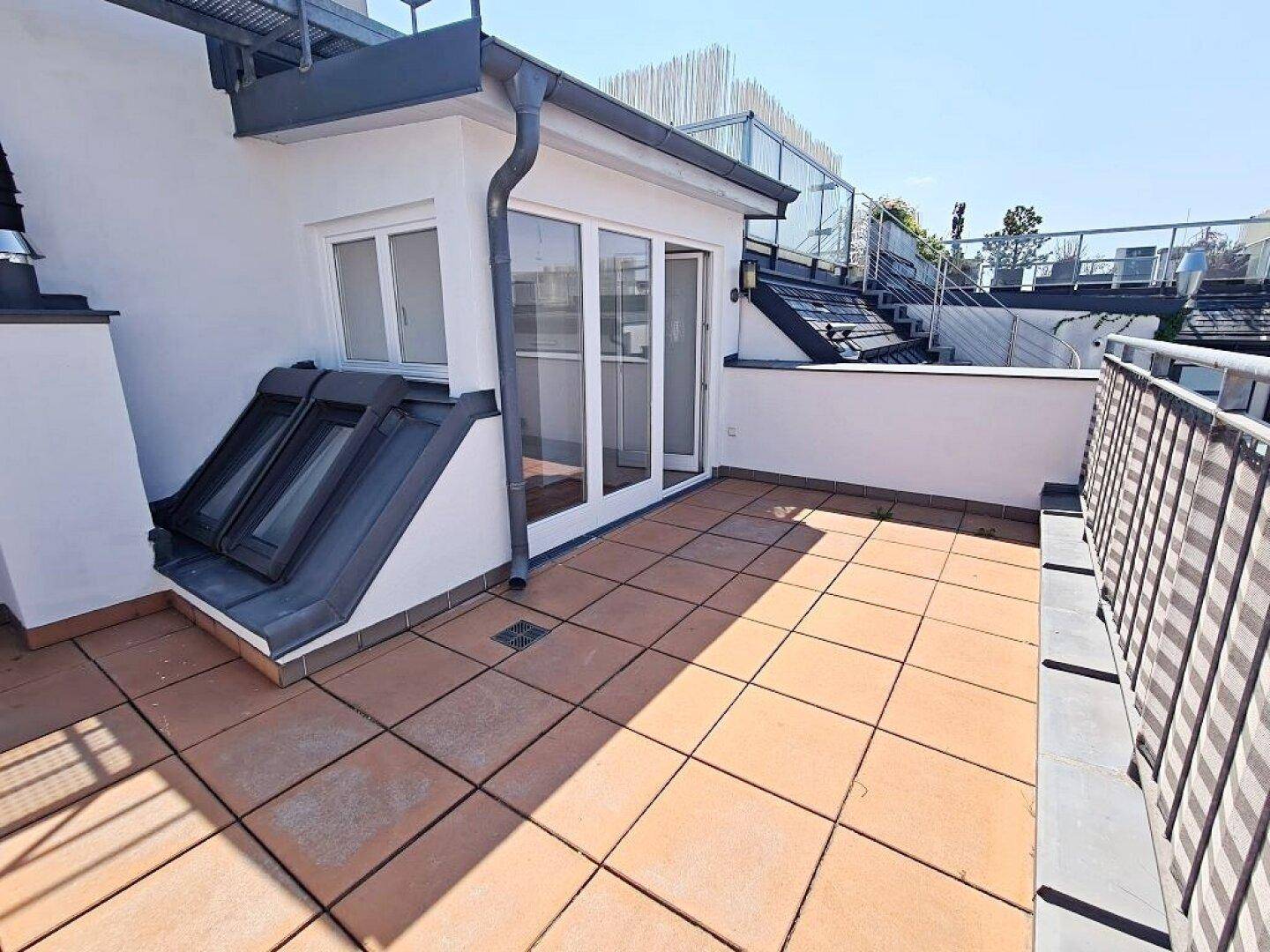 Weitläufige Dachterrasse mit Fliesenboden, umgeben von weißer Fassade und Dachfenstern, sehr hell.
