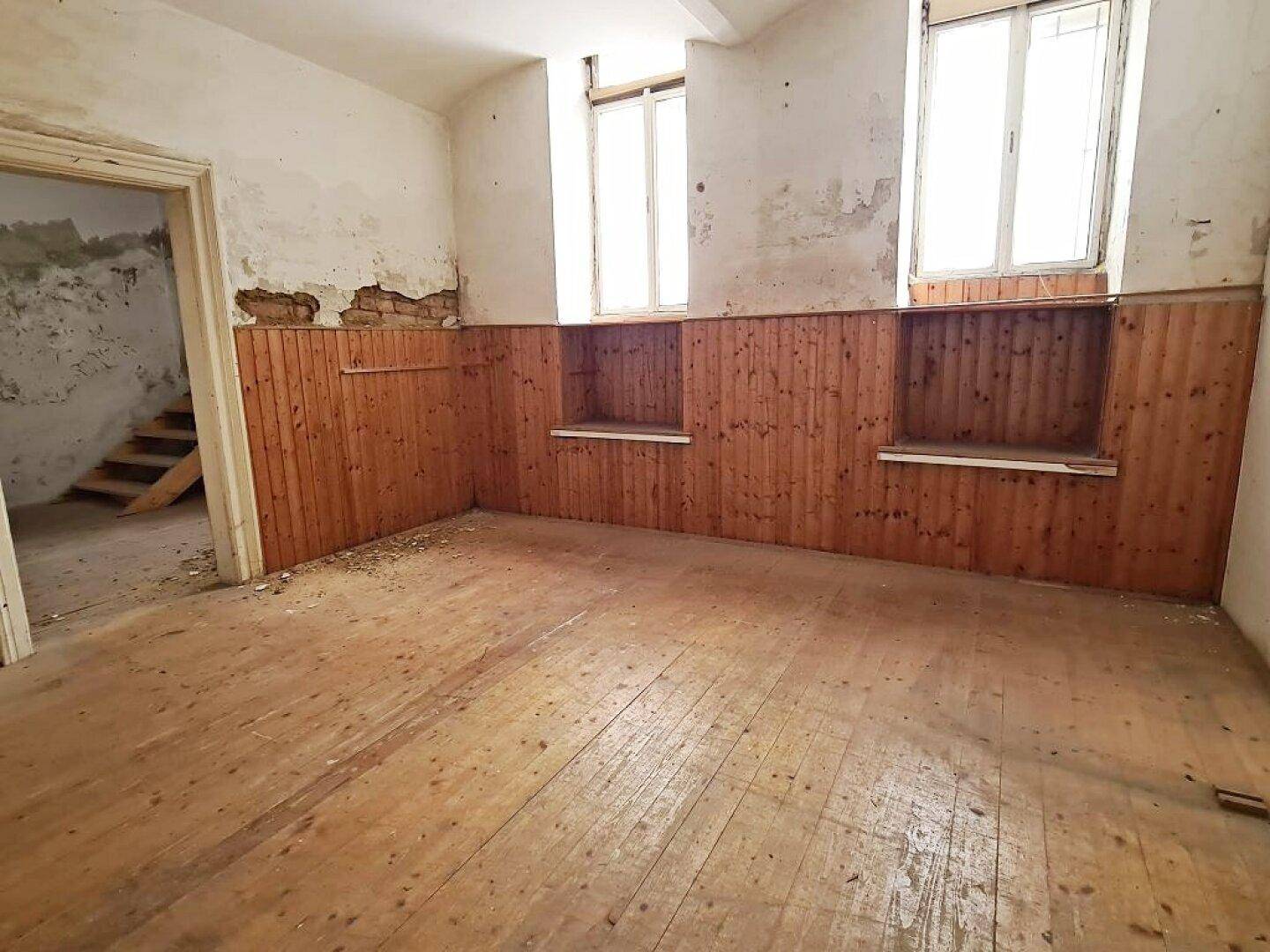 Leerer Raum mit Holzdielenboden, Holzvertäfelung an den Wänden und zwei Fenstern, die Tageslicht hereinlassen.
