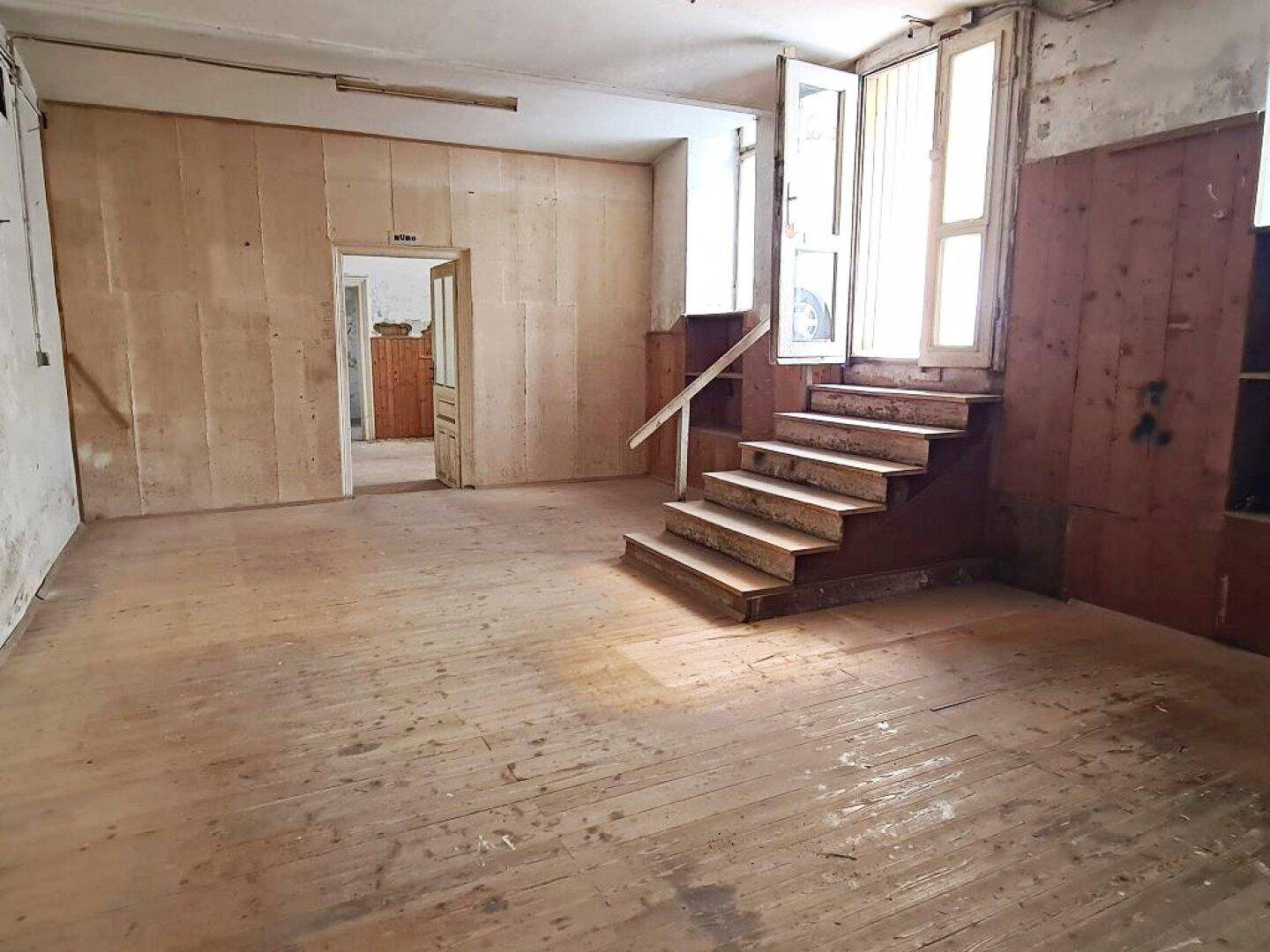 Geräumiger, leerer Innenraum mit Holzdielenboden, einer Holztreppe und einem Durchgang zu einem weiteren Zimmer.
