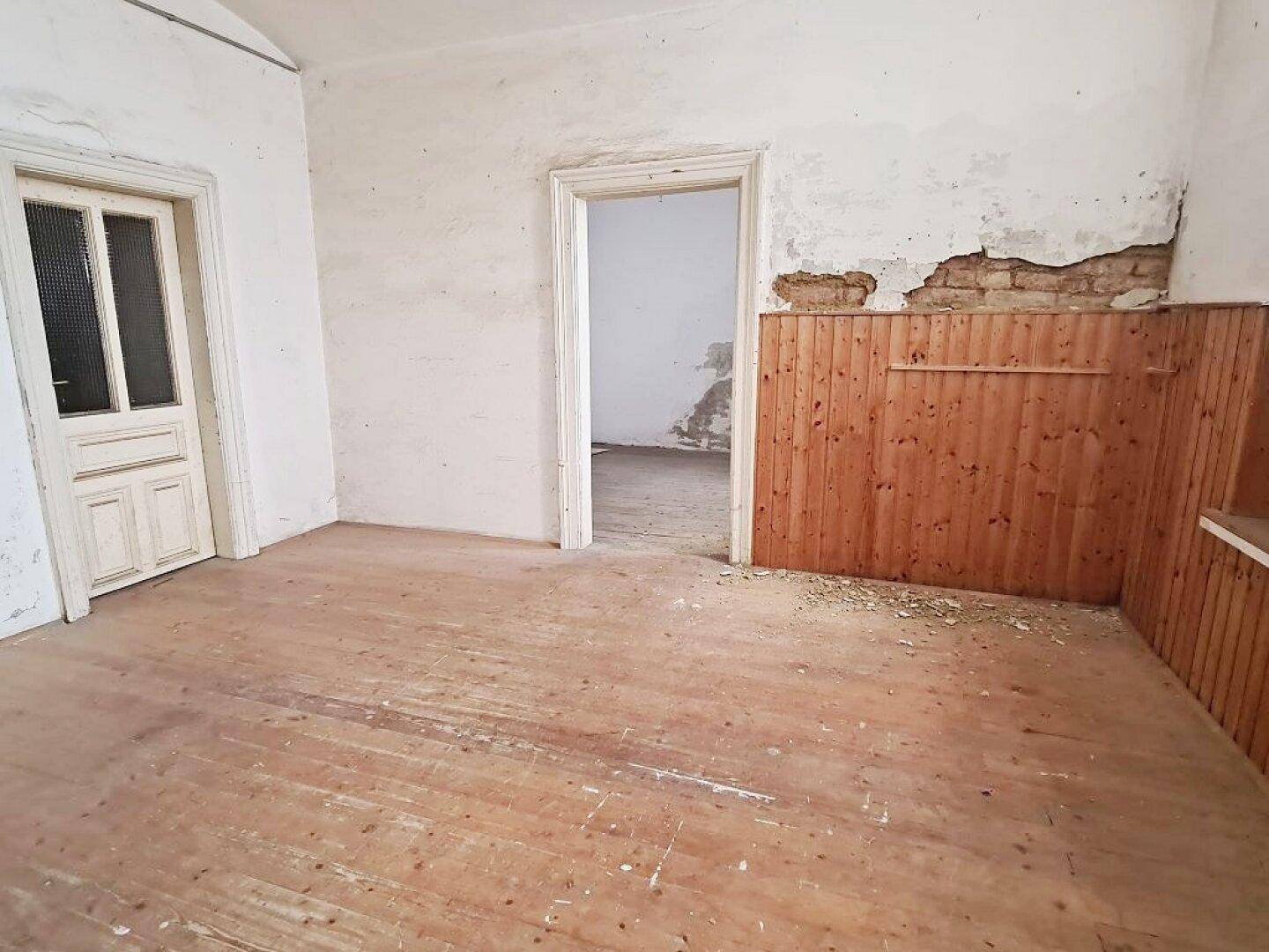 Leerer Raum mit Holzdielenboden, einer weißen Tür und einem offenen Durchgang zu einem weiteren Zimmer.