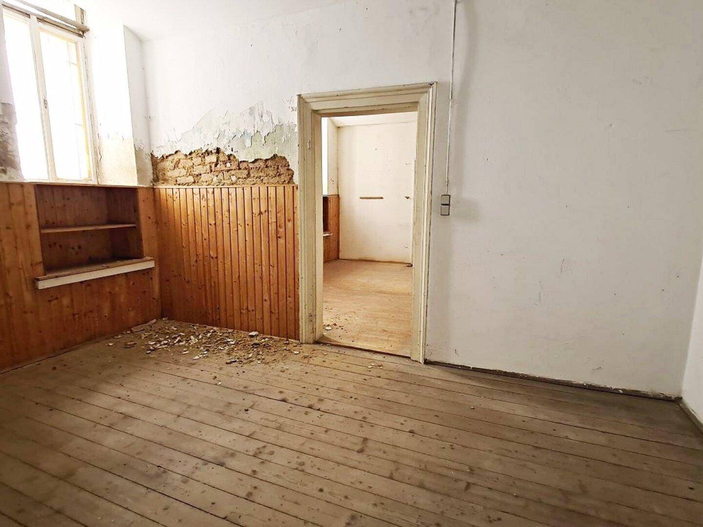 Leerer Raum mit Holzdielenboden, einem offenen Durchgang zu einem weiteren Zimmer und Holzvertäfelung an einer Wand.