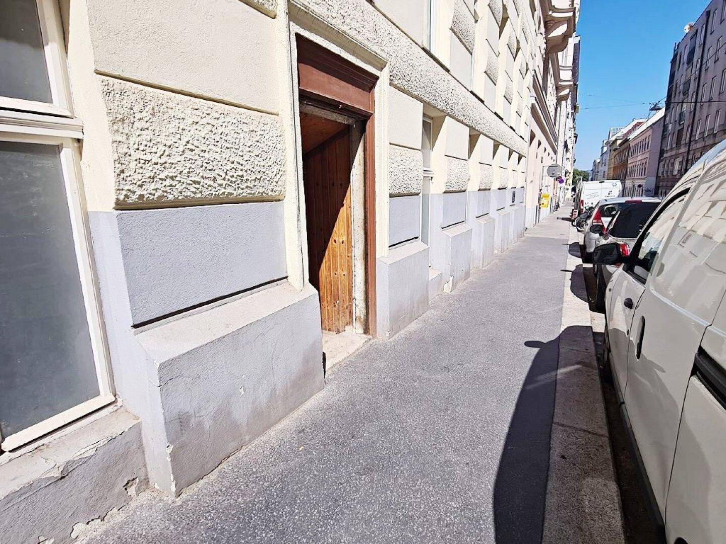 Eingangsbereich eines Gebäudes von der Straße aus gesehen, mit Bürgersteig und geparkten Autos.