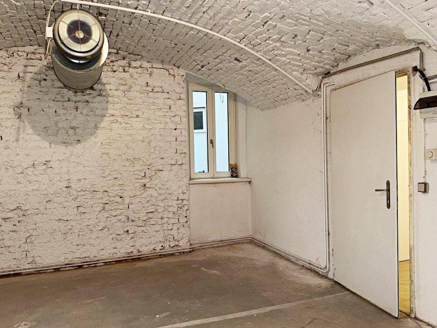 Gewölbter Kellerraum mit Fenster, weiß gestrichenen Ziegelwänden und einem Lüftungsgerät an der Decke.