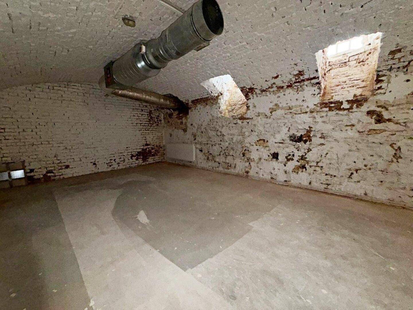 Kellerraum mit freiliegenden Ziegelwänden, kleinen Fenstern und einem großen Lüftungsrohr an der gewölbten Decke.