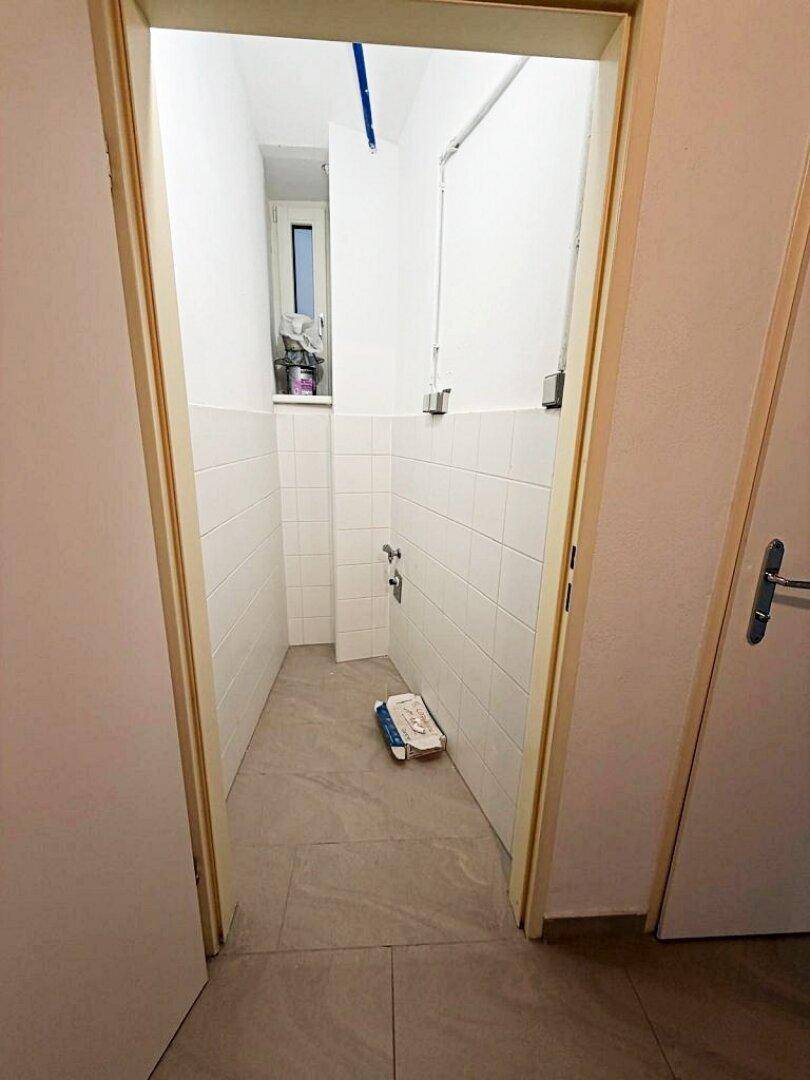 Schmaler Raum mit weißen Wandfliesen und grauen Bodenfliesen, möglicherweise für eine Dusche oder als Abstellraum.