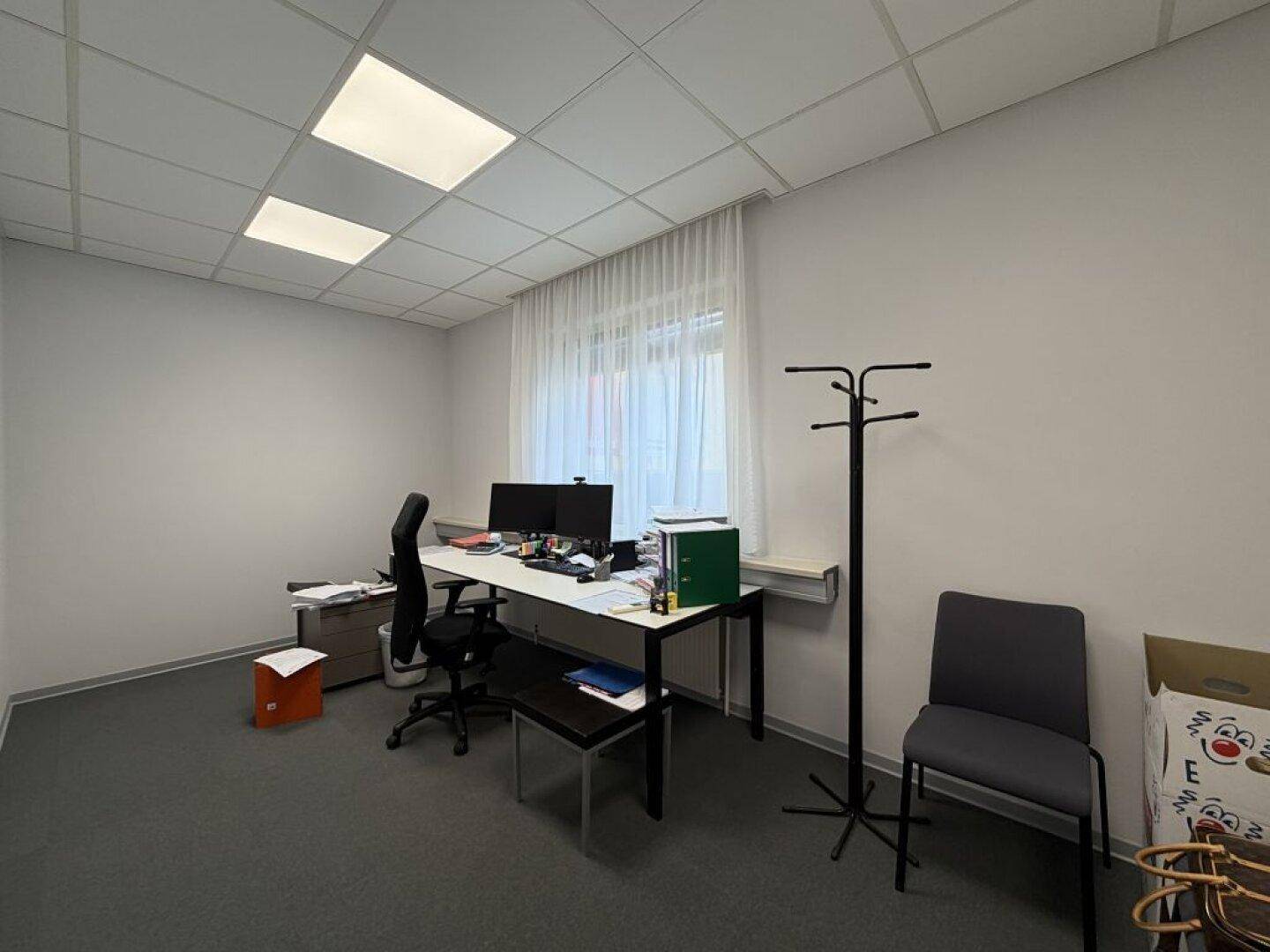 Einzelbüro mit Schreibtisch, Bürostuhl, Teppichboden und einem Fenster mit Vorhängen.