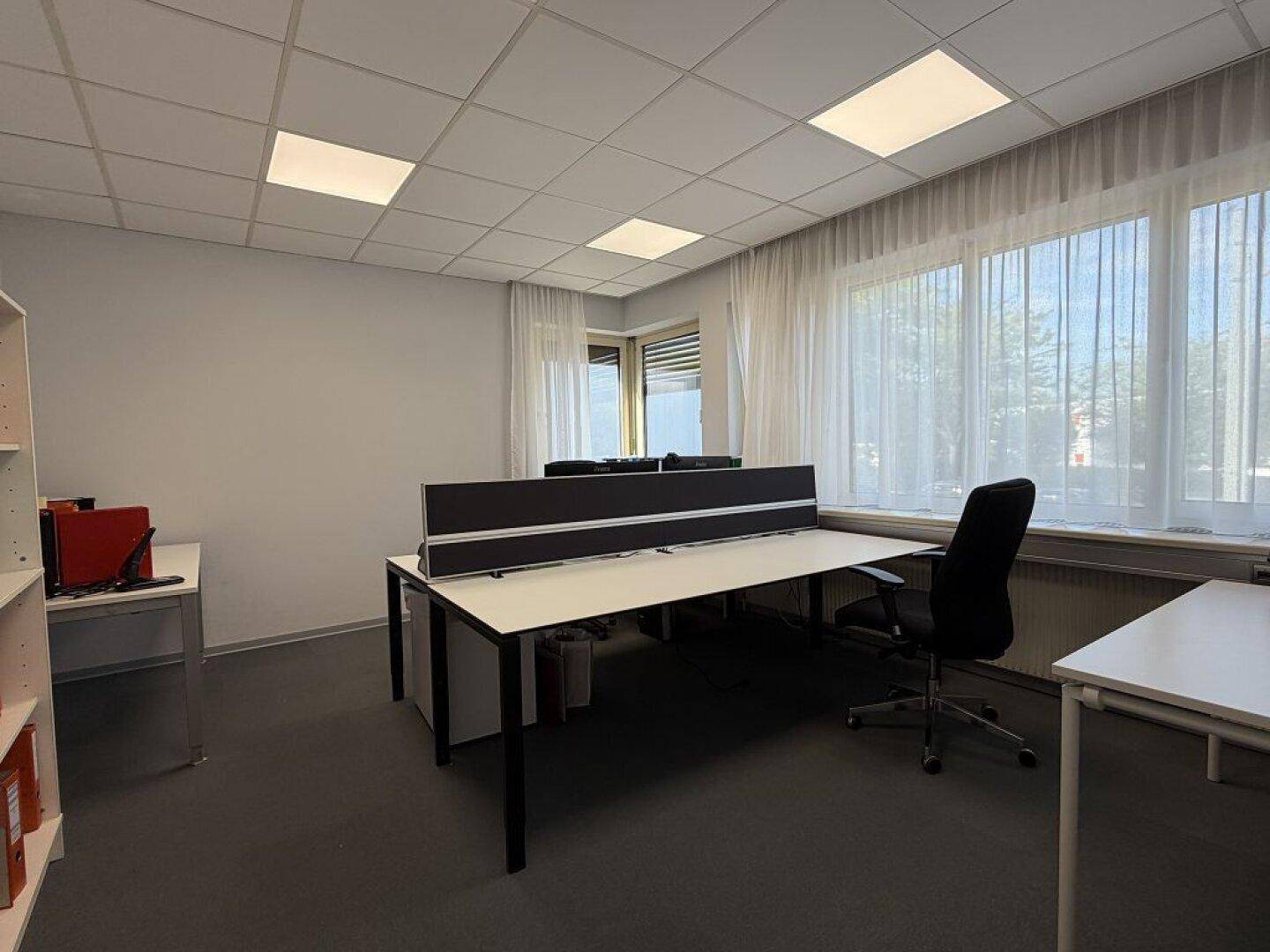 Büro mit mehreren leeren Schreibtischen, Teppichboden und großen Fenstern mit Vorhängen.