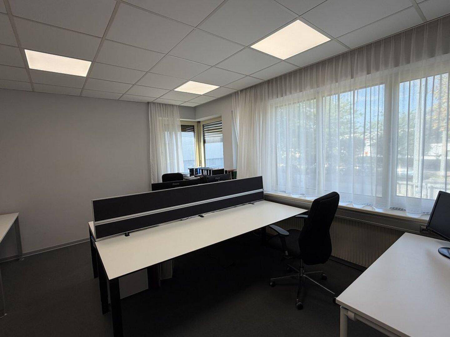 Büro mit mehreren Arbeitsplätzen, Teppichboden und großen Fenstern, die viel Tageslicht hereinlassen.