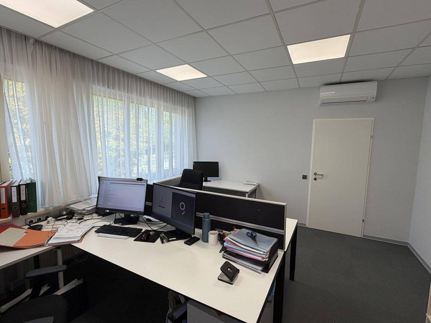 Büroarbeitsplatz mit zwei Monitoren, Aktenordnern und einem Klimagerät an der Wand.