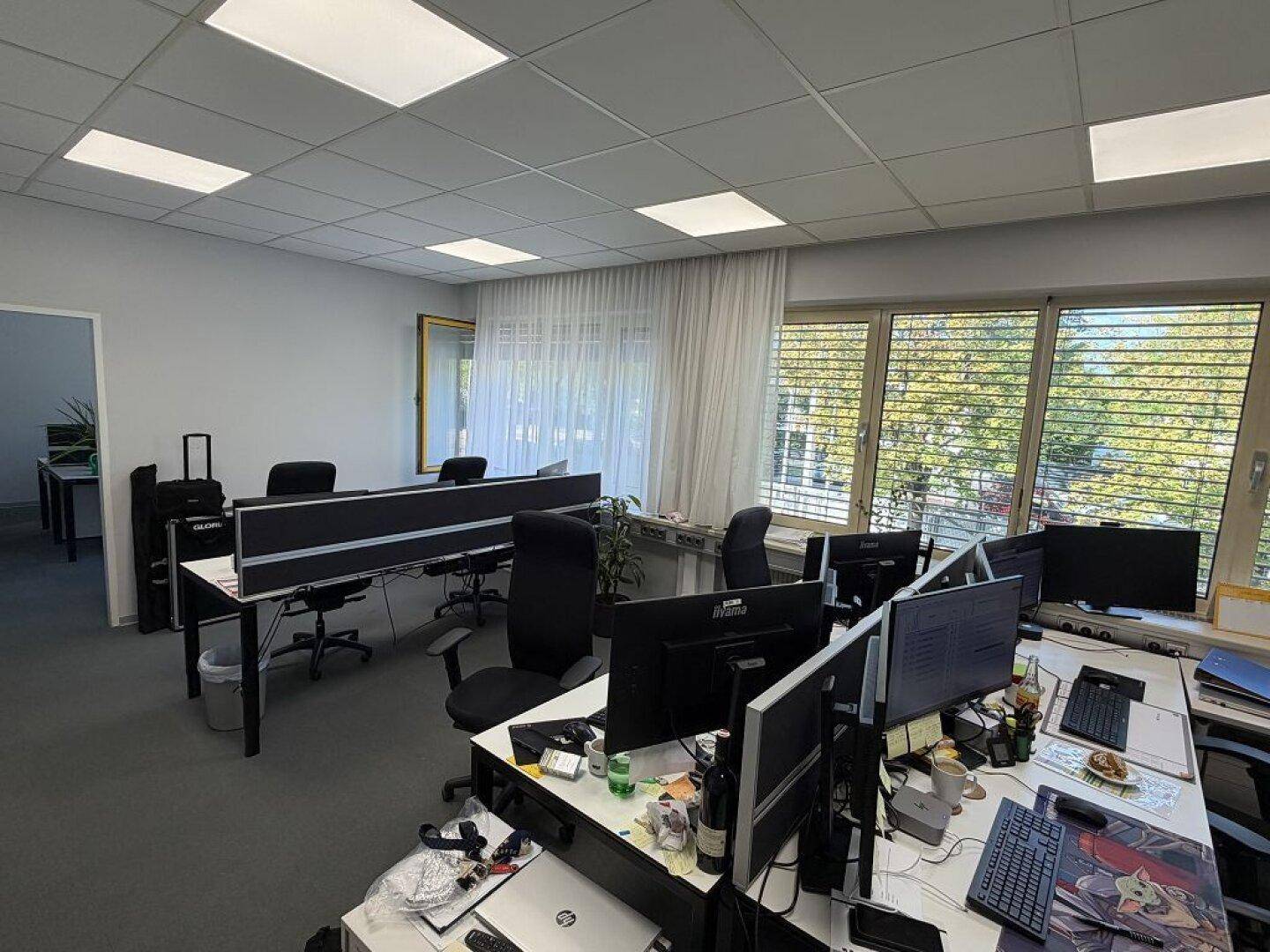 Großraumbüro mit mehreren Arbeitsplätzen, Teppichboden und großen Fenstern, die viel Tageslicht hereinlassen.
