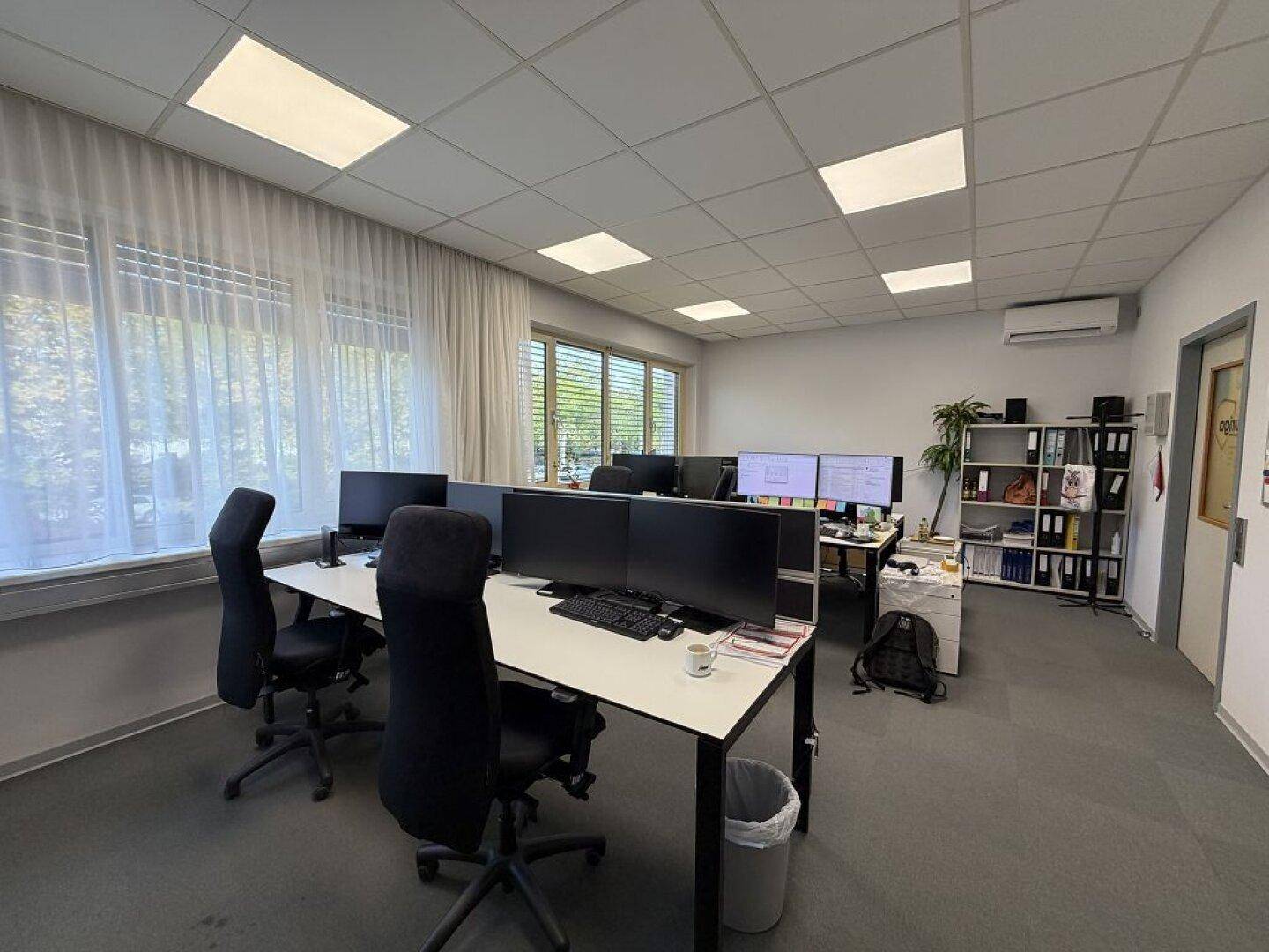 Großraumbüro mit mehreren Arbeitsplätzen, Teppichboden, Klimaanlage und Regalen an der Wand.