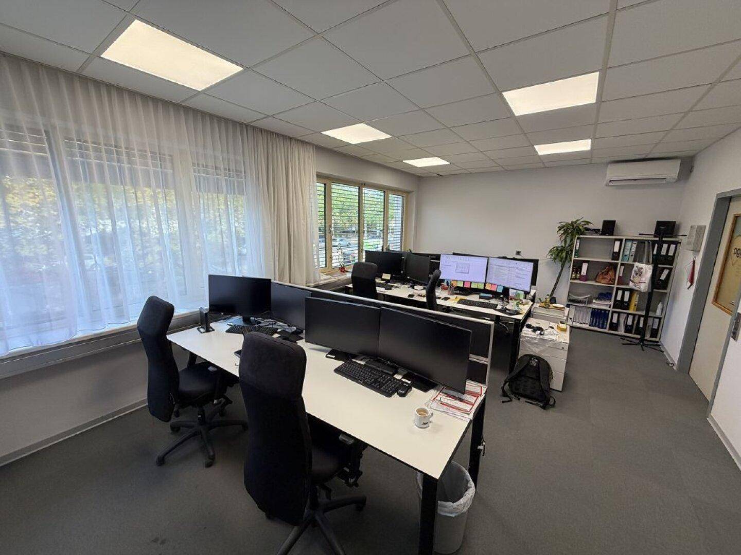 Großraumbüro mit mehreren Arbeitsplätzen, Teppichboden, Klimaanlage und Regalen an der Wand.