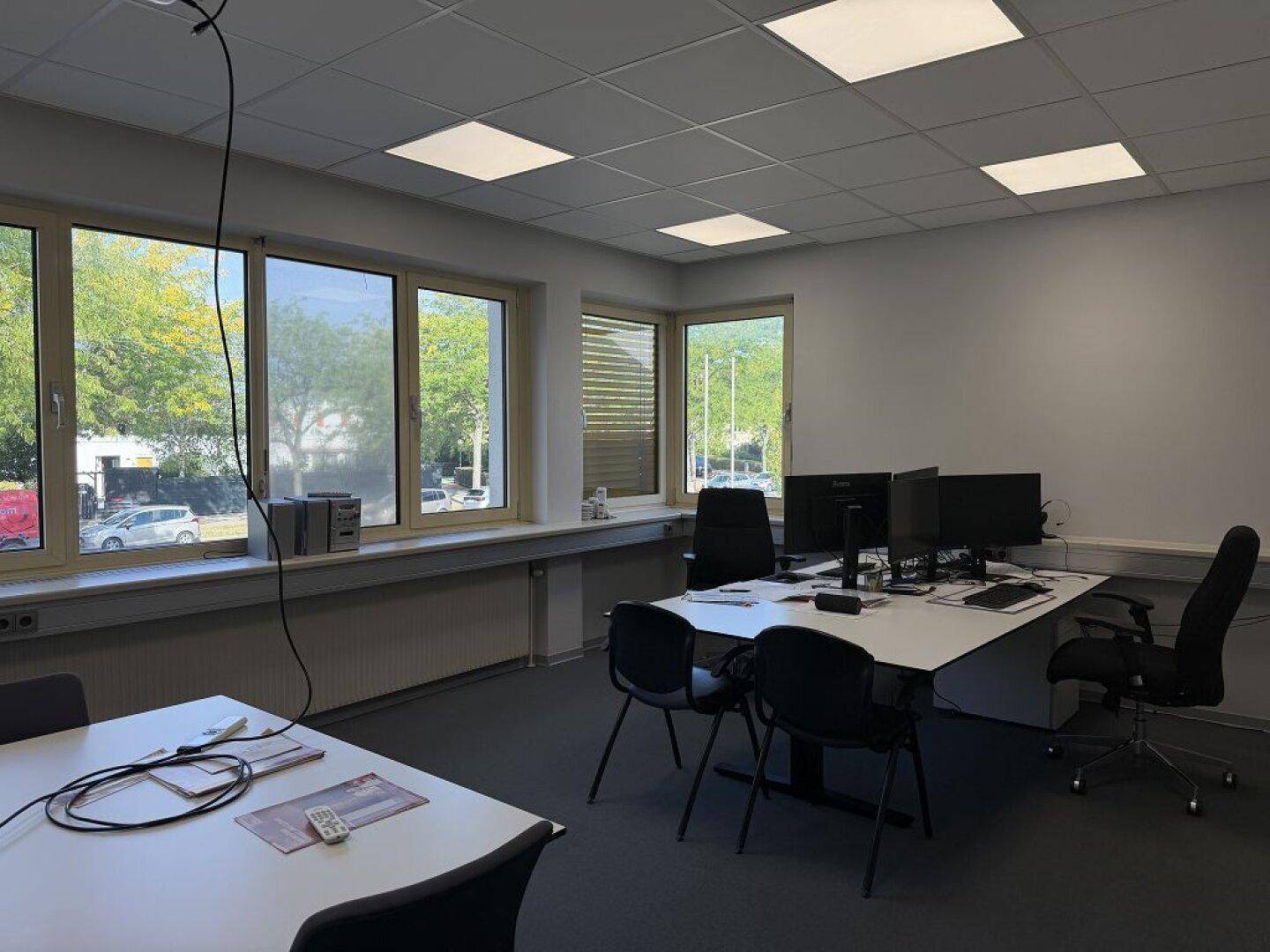 Büroraum mit mehreren Arbeitsplätzen, Teppichboden und großen Fenstern mit Blick ins Grüne.
