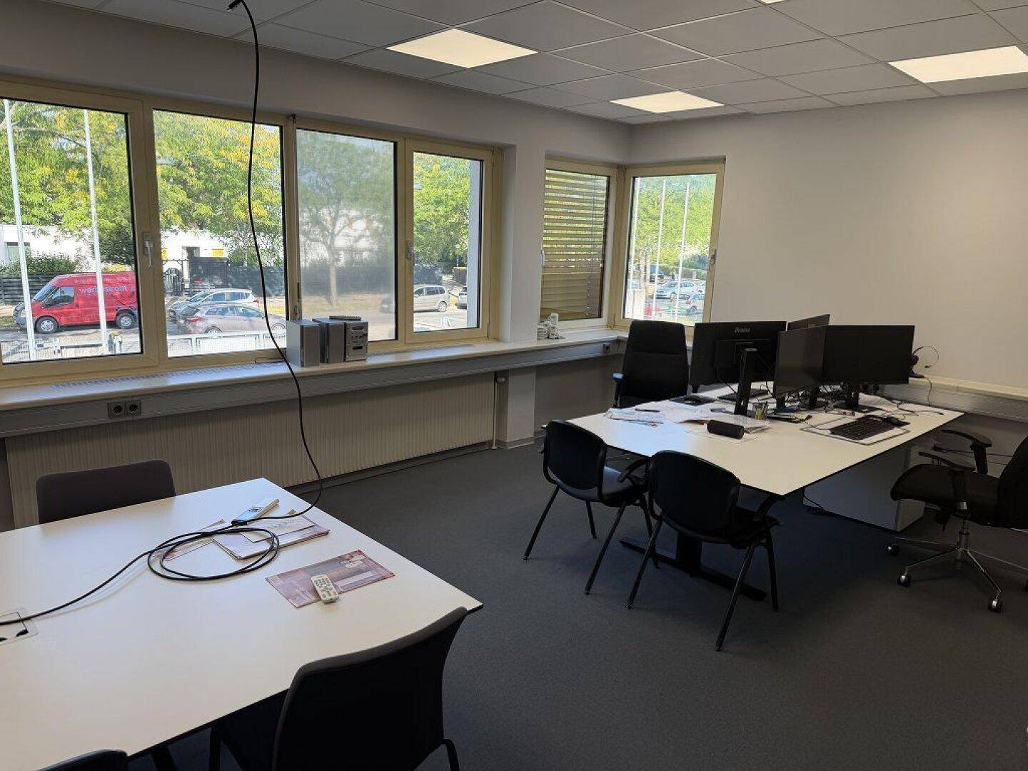 Büroraum mit mehreren Arbeitsplätzen, Teppichboden und großen Fenstern mit Blick ins Grüne.