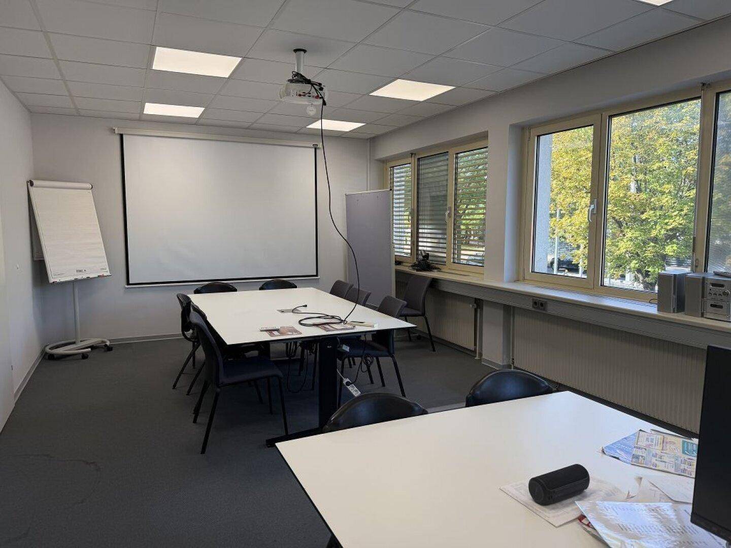 Konferenzraum mit großem Tisch, Stühlen, Beamer und Leinwand, sowie Fenstern mit Blick ins Grüne.