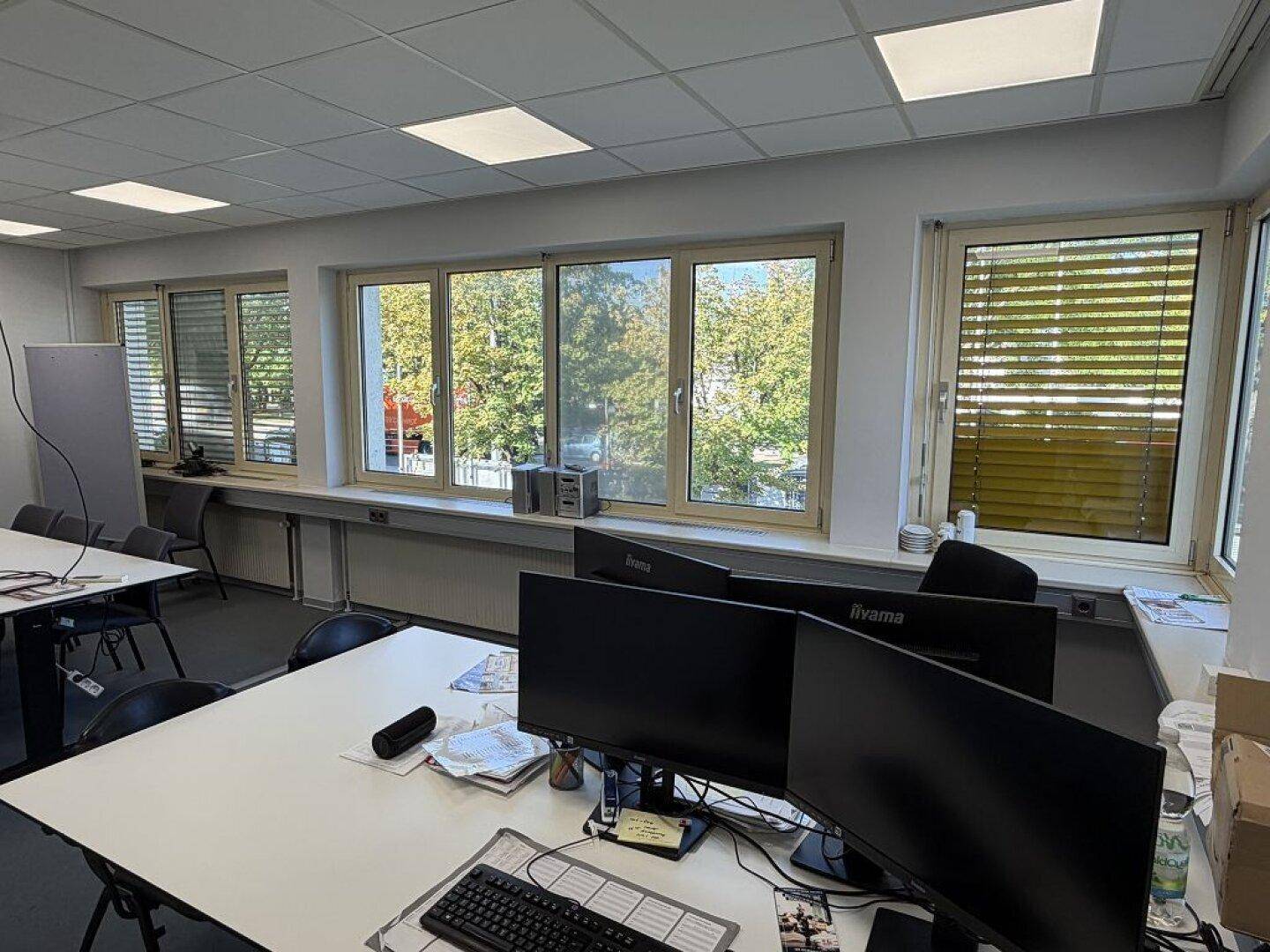 Büroraum mit mehreren Arbeitsplätzen, Teppichboden und großen Fenstern mit Jalousien.