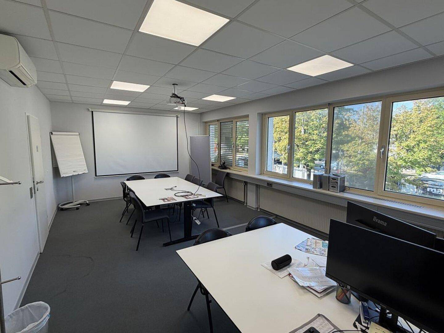 Geräumiger Büroraum mit Konferenztisch, Projektor und großen Fenstern, die viel Tageslicht hereinlassen.
