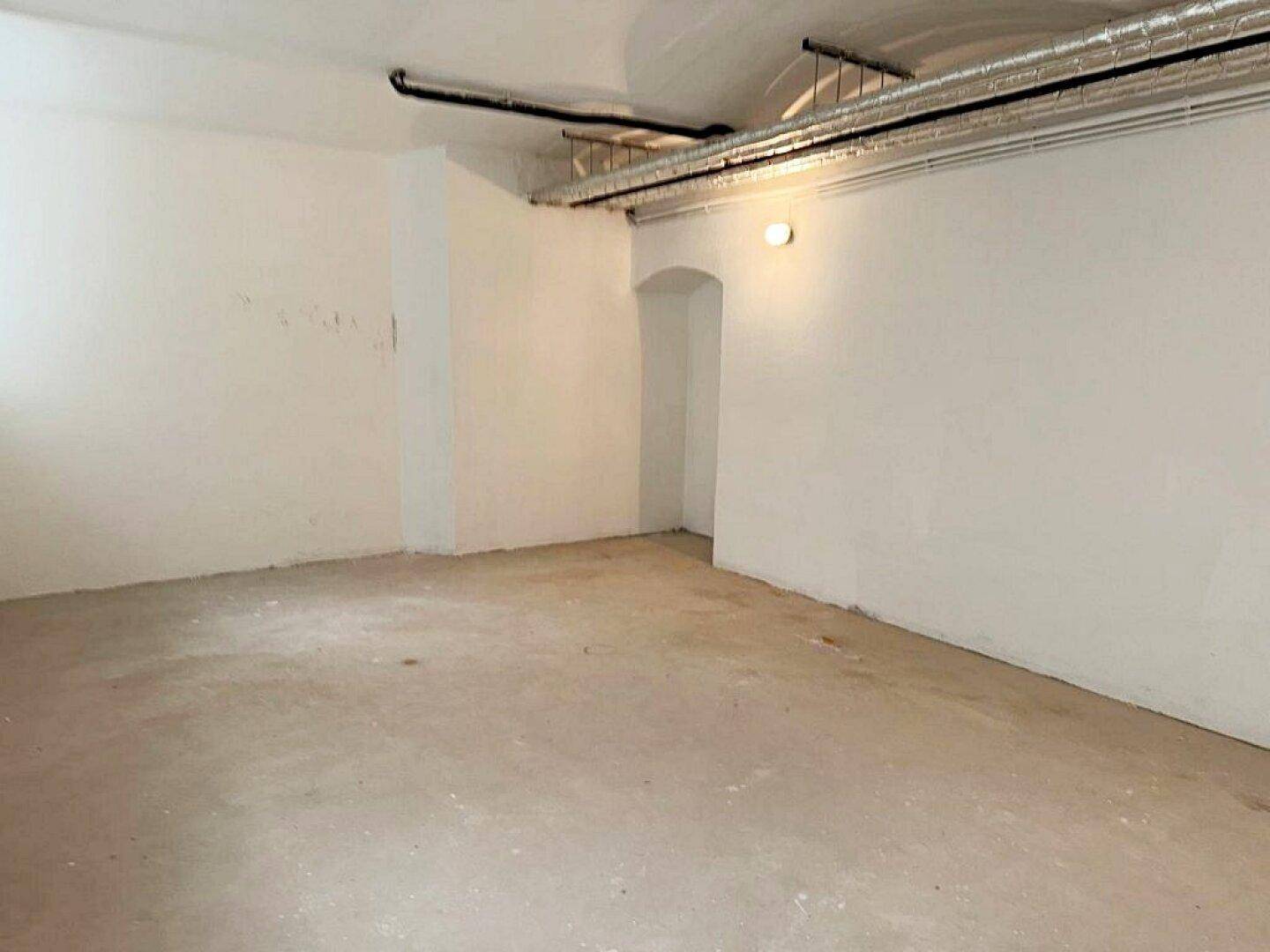 Unfertiger Kellerraum mit Betonboden, weißen Wänden, sichtbaren Deckenleitungen und einem gewölbten Durchgang.
